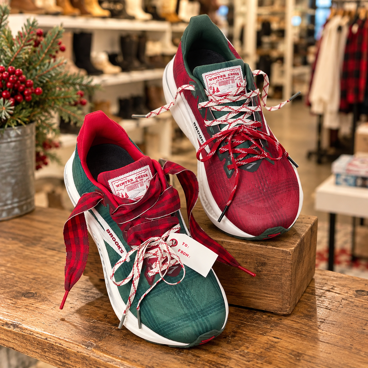 Brooks Ghost 16 Winter Cheer Christmas running shoes featuring one red shoe and one green shoe, both with festive holiday plaid accents and decorative laces, displayed on a rustic wooden table inside a cozy shoe boutique with Christmas greenery and softly blurred store shelves in the background.